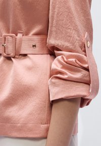 Ροζ, ζαρωτό μπλούζα με ζώνη, που διαθέτει μια μεγάλη ορθογώνια αγκράφα και μανίκια που έχουν γυριστεί και στερεωθεί με κουμπιά. Μαλακή υφή, καθημερινό σχέδιο.