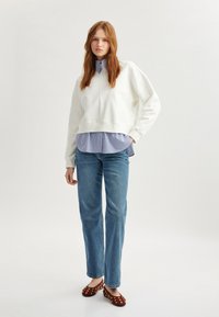 Biała krótka bluza, niebieska koszula w paski z kołnierzykiem pod spodem, niebieskie dżinsy i brązowe buty w kropki. Casualowy styl.