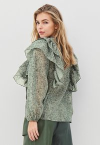 Grön, lätt blus med volangdetaljer längs axlarna och ryggen. Texturerat tyg med subtilt mönster; långa ärmar med elastiska manschetter.