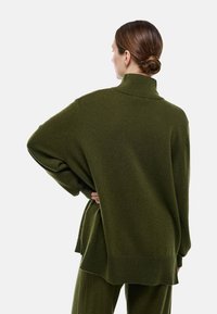 Pull en tricot oversize vert foncé avec un col montant, des épaules tombantes et des poignets côtelés ; texture douce avec une coupe décontractée et des fentes latérales.
