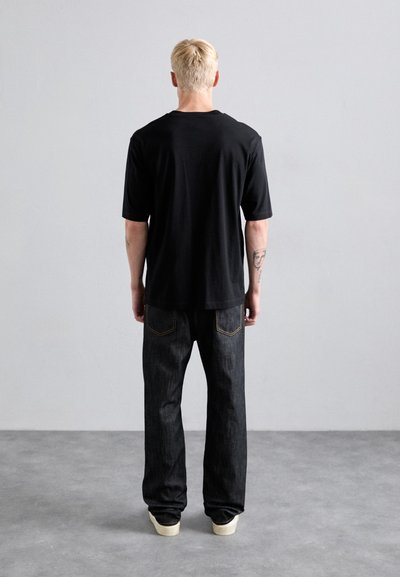 Personne blonde avec des tatouages, portant une chemise noire à manches courtes, un jean sombre et des chaussures blanches, se tenant face à un mur gris clair uni.