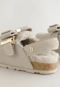 Sandales pour enfants de couleur beige clair avec un intérieur en peluche, des nœuds décoratifs et des accents dorés, dotées d'une semelle en liège pour plus de confort.