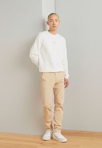 Hanorac alb cu un design minimalist și pantaloni chinos bej deschis. Încălțămintea este formată din teniși albi. Modelul stă în fața unui fundal neutru.