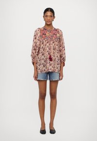Blusenshirt mit floralem Druck und weiten Ärmeln, aus leichtem Stoff in Rot- und Blautönen, mit einem Schlüsselloch-Ausschnitt und Quasten-Details. Kombiniert mit Jeans-Shorts.