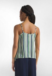 Kobieta z warkoczami, ubrana w bezrękawnik w pionowe paski w kolorach niebieskim, zielonym i białym oraz granatowe spodnie, widziana od tyłu.