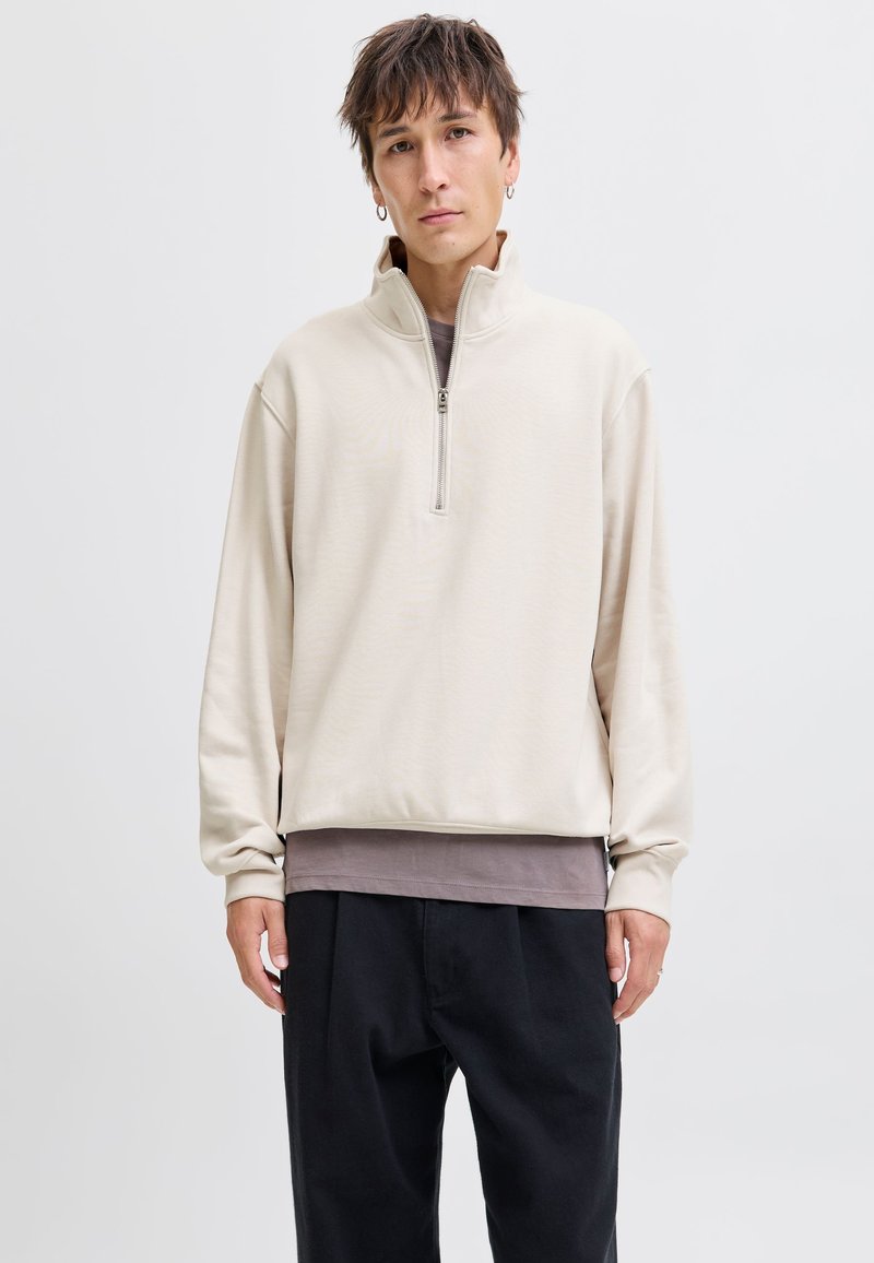 Młoda osoba z krótkimi brązowymi włosami, ubrana w beżową bluzę z krótkim zamkiem, czarne spodnie i kolczyki koła, stojąca na tle jednolitego białego tła.