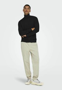 Man staat met zijn armen gekruist, draagt een zwarte coltrui, beige broek en witte sneakers voor een effen lichte achtergrond.