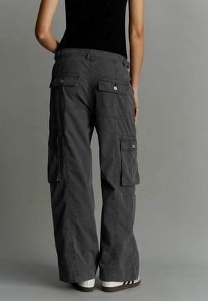 Persoon draagt losse donkergrijze cargobroek met klepzakken en witte sneakers, staand tegen een effen grijze achtergrond.