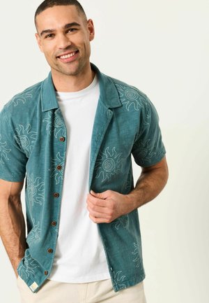 Lächelnder Mann, der ein türkisfarbenes kurzärmeliges Hemd mit weißen Sonnenmustern über einem weißen T-Shirt trägt und das Hemd mit einer Hand offen hält.