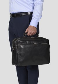 Sorte læder aktetaske med dobbelte håndtag, to lynlåse og en glat tekstur. Båret af en person i formelt tøj.