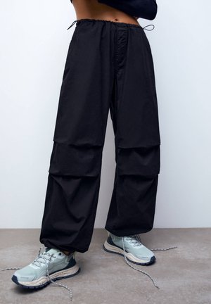 Persona con pantalones cargo negros holgados con cintura elástica y zapatillas gris claro con cordones desatados, de pie sobre un suelo gris.