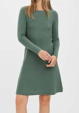 Femme portant une robe longue en maille verte à manches longues et longueur genou, avec un col rond, debout les mains légèrement jointes.