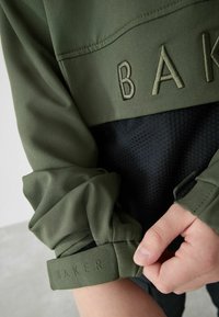 Giacca verde oliva con pannello nero testurizzato, caratterizzata dal logo in rilievo "BAKER" sul polsino della manica e regolazione in velcro sul polsino.