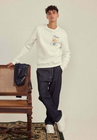 Sweatshirt blanc avec un design de poche florale, associé à un pantalon en denim foncé. L'arrière-plan présente une chaise en bois et un tapis à motifs. Baskets blanches.