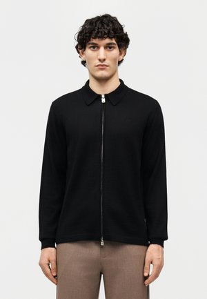 Νεαρός άνδρας με καστανά σγουρά μαλλιά φοράει μαύρο πουλόβερ με φερμουάρ και γιακά και μπεζ παντελόνι, στέκεται μπροστά σε απλό φωτεινό φόντο.