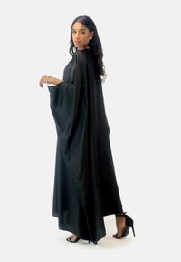 Robe cape en soie noire, longue jusqu'au sol, avec des manches larges. Associée à des sandales noires à talons hauts. Silhouette simple et élégante avec des accents minimaux.