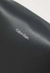 Juodas odos paviršius su tekstūruotais raštais, kuriame aiškiai matomas baltas spausdintas logotipas „Calvin Klein“. 