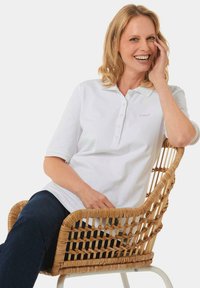 Weißes Polohemd mit einem Knopfkragen und kurzen Ärmeln, mit einer dezenten Logo-Stickerei, kombiniert mit dunklen Jeans, während er in einem geflochtenen Stuhl sitzt.