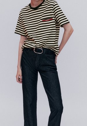 Personne portant une chemise à manches courtes rayée noir et blanc avec un détail de poche rouge, une ceinture noire et un jean droit foné sur un fond uni.