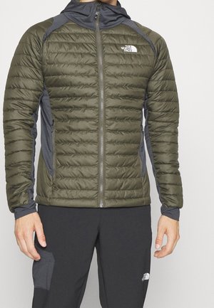 Homme portant une doudoune matelassée vert olive et grise avec une fermeture éclair avant, et un pantalon noir avec une poche latérale, tous deux de la marque The North Face.