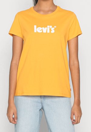 Kvinde iført en gul Levi's t-shirt med hvidt logo tekst og lyseblå jeans, vist fra nakke til hofter mod en ensfarvet baggrund.