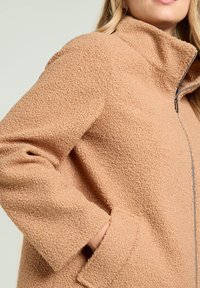 Giacca in pile testurizzato beige con colletto alto, zip frontale completa e tasche laterali, caratterizzata da un aspetto morbido e accogliente.