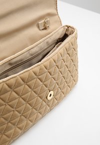 Beżowa pikowana torba o miękkiej fakturze, wyposażona w złote zapięcie na zatrzask i wewnętrzną kieszonkę. Całość ozdobiona eleganckim kwadratowym wzorem.