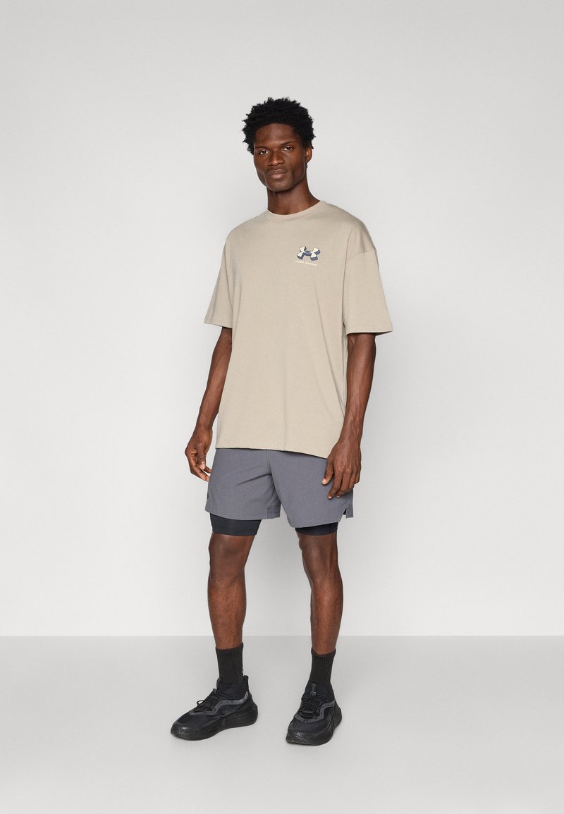 T-shirt in cotone beige con un piccolo logo sul petto, abbinato a pantaloni corti grigio scuro. Il modello indossa sneakers nere e calzini alla caviglia.