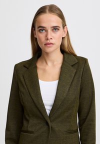 Olivengrüner Herzchenblazer mit Revers, zwei Fronttaschen und tailliertem Design, getragen über einem weißen Oberteil.