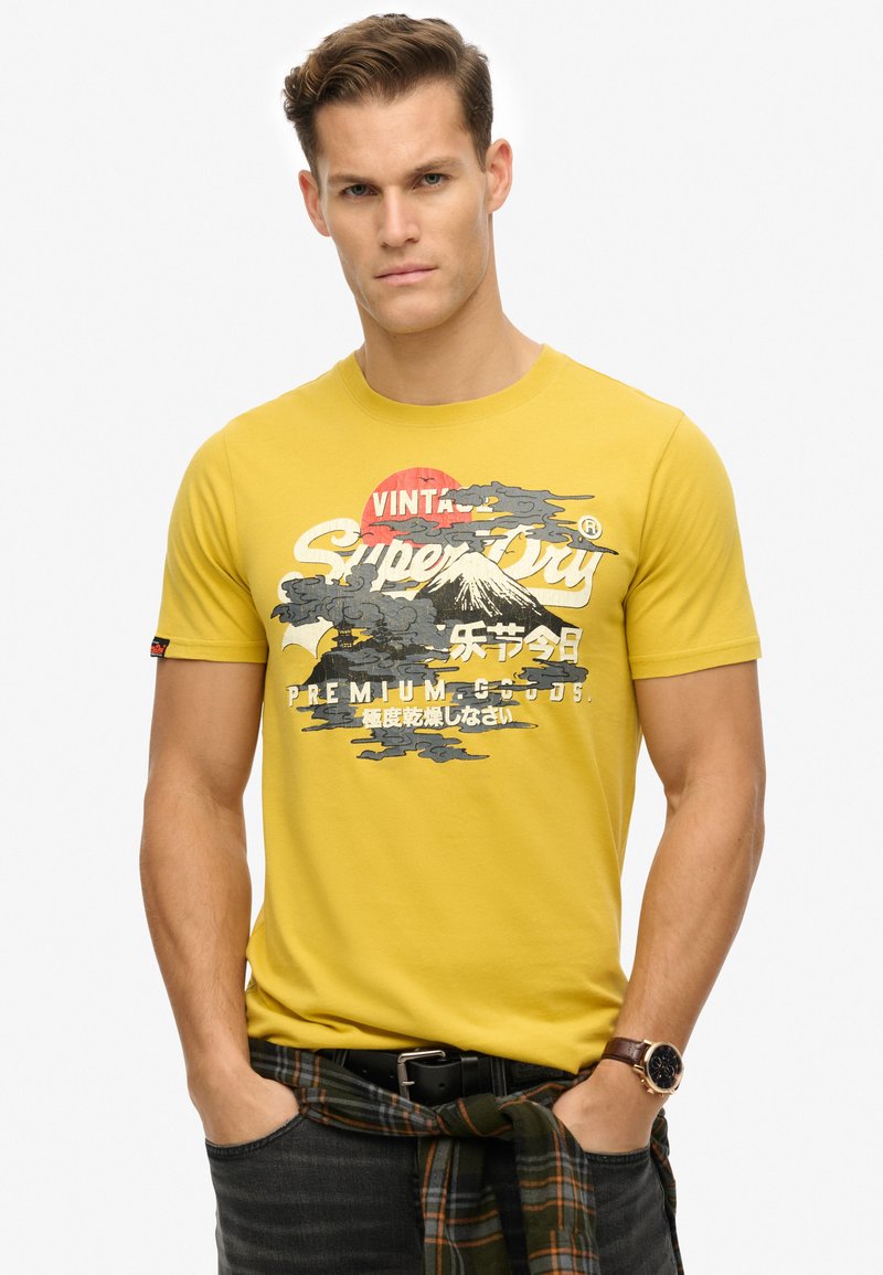 Camiseta de algodón amarilla con un estampado gráfico de montaña y sol, de mangas cortas, cuello redondo y una etiqueta con el logo en el lateral.