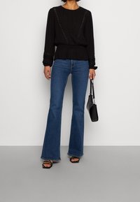 Musta pitkähihainen paita, jossa on tekstuurista kangasta ja koristeellisia ompeleita, yhdistettynä sinisiin leveälahkeisiin farkkuihin ja mustiin remmisandaaleihin, pitäen mustaa käsilaukkua.