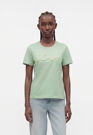 Femme aux cheveux tressés portant un T-shirt vert clair avec le mot « Love » en lettres perlées et un jean bleu clair, debout sur un fond blanc.