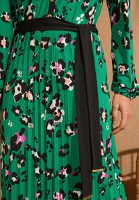 Abito plissettato verde con cintura nera, caratterizzato da un motivo animalier leopardato in bianco, nero, rosa e dettagli verdi. Dettaglio in metallo dorato sulla cintura.