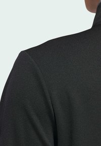 Veste noire texturée avec un col haut, présentant un léger éclat et une finition lisse. Le design comprend une couture d'épaule structurée.