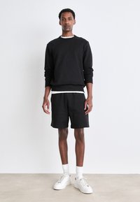 Svart bomullsgenser med ribbestrikkede mansjetter, sammen med svarte shorts. Modellen har på seg hvite sneakers og ankelsokker. Enkelt, uformelt design.