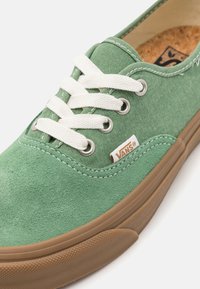 Baskets en daim vert avec lacets blancs, semelle en caoutchouc marron et œillets métalliques. Présente une étiquette du logo Vans sur le côté et une surface texturée.