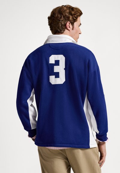 Muž ve modrobílém tričku s dlouhým rukávem s bílým číslem 3 na zádech, béžové kalhoty, stojící bokem na jednobarevném pozadí.