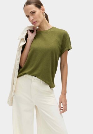 Kvinde iført olivengrøn t-shirt og cremefarvede bukser med vide ben, der holder en lys jakke over skulderen mod en ensfarvet baggrund.