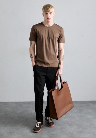 Barna rövid ujjú póló, fekete nadrág, sötét cipők világos fűzővel, nagy barna bőr táska a kezében. Egyszerű szürke háttér.