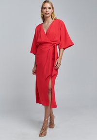 Rotes Wickelkleid mit weiten Ärmeln, tiefem V-Ausschnitt, seitlichem Schlitz und Bindegürtel. Kombiniert mit goldenen Riemchensandalen. Glattes, fließendes Material.