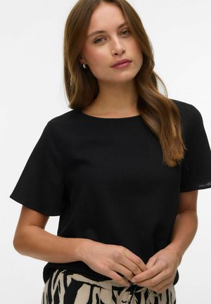 Jeune femme aux longs cheveux bruns portant un haut noir à manches courtes et un pantalon à motifs clairs et foncés, debout avec les mains jointes.