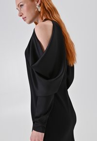Schwarzes Kleid mit drapiertem, schulterfreiem Design, langen Ärmeln und glattem, glänzendem Stoff. Sichtbares silbernes Ohrringdetail.