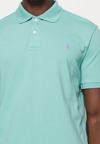 Polo triko v mátové zelené barvě vyrobené z bavlny. Má klasický límec, dvě knoflíkové légy a malé fialové vyšívané logo na hrudi.