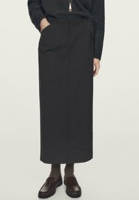 Woman wearing a black long skirt with pockets, dark sweater, brown socks, and dark brown slip-on loafers, standing with one hand in pocket.