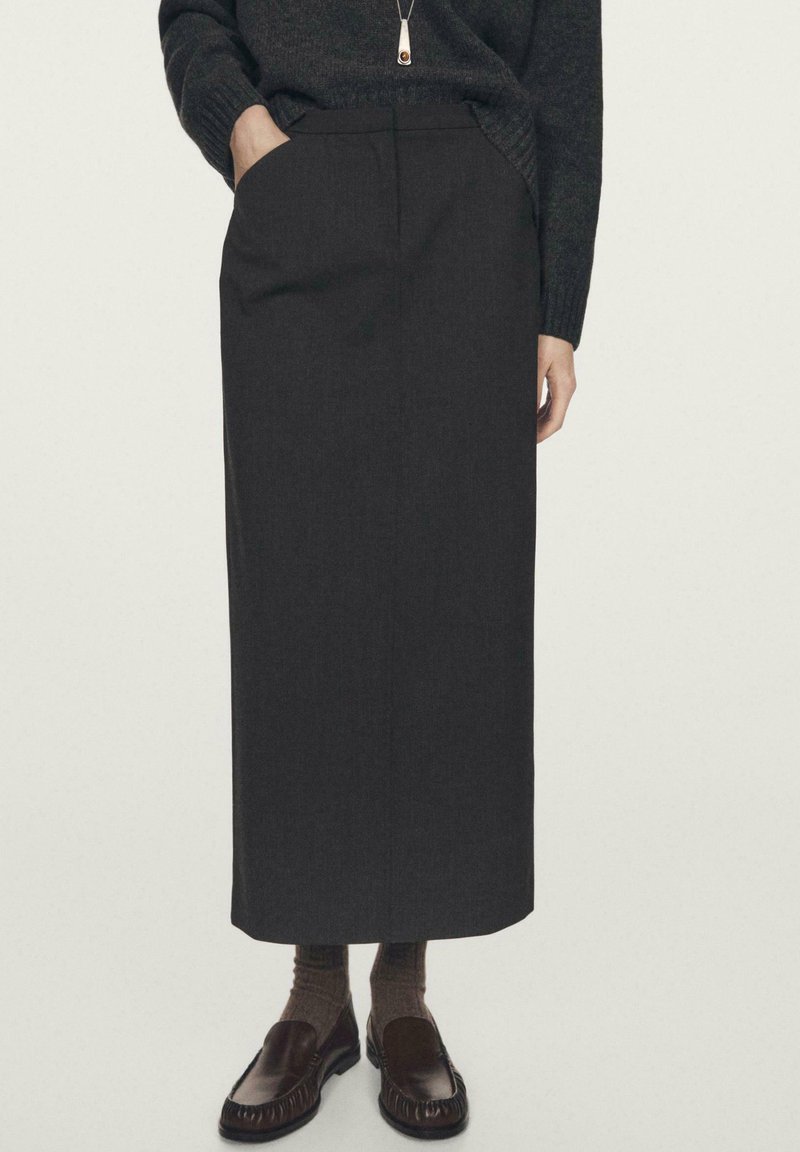 Woman wearing a black long skirt with pockets, dark sweater, brown socks, and dark brown slip-on loafers, standing with one hand in pocket.