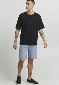 T-shirt en coton noir à manches courtes, associé à un short en denim bleu clair. Le modèle porte des baskets noires et a des tatouages visibles sur les bras.