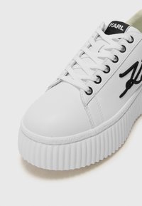 Hvite plattform sneakers laget av glatt syntetisk materiale, med rundt tå, snøring, svart logo og strukturert gummisåle med detaljer.