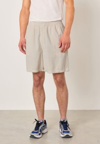 Mężczyzna w beżowych sztruksowych szortach i niebiesko-szarych butach do biegania, stojący na jasnej, gładkiej podłodze na tle białej ściany.