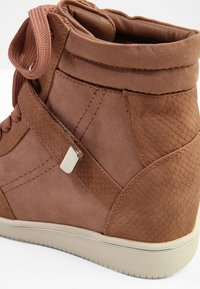 Hnědý teniskový kotník s texturovanými panely, semišovými detaily, tlustou podrážkou, polstrovaným límcem a hnědými tkaničkami ve stejné barvě.