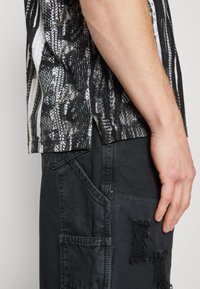 Chemise à manches courtes avec un motif abstrait noir et blanc, associée à un short cargo gris foncé avec des déchirures et de multiples poches.